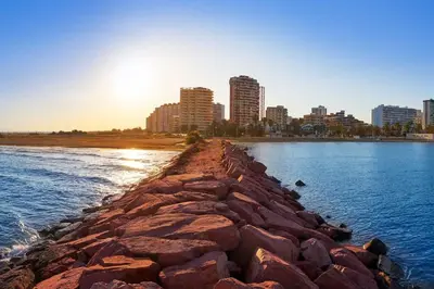 a rock wall in the water with a city in the background at La Llar45 , Relájate junto al mar in Playa Pobla de Farnals