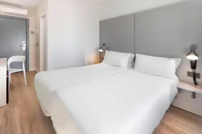 a white bedroom with a large white bed and a table at B&B HOTEL Valencia Aeropuerto in Paterna