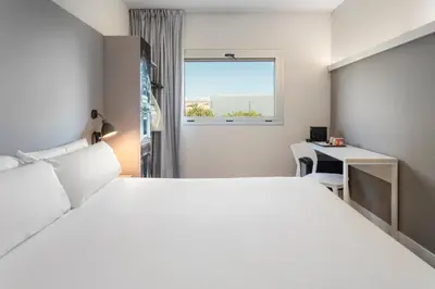 a bedroom with a white bed and a desk and a window at B&B HOTEL Valencia Aeropuerto in Paterna